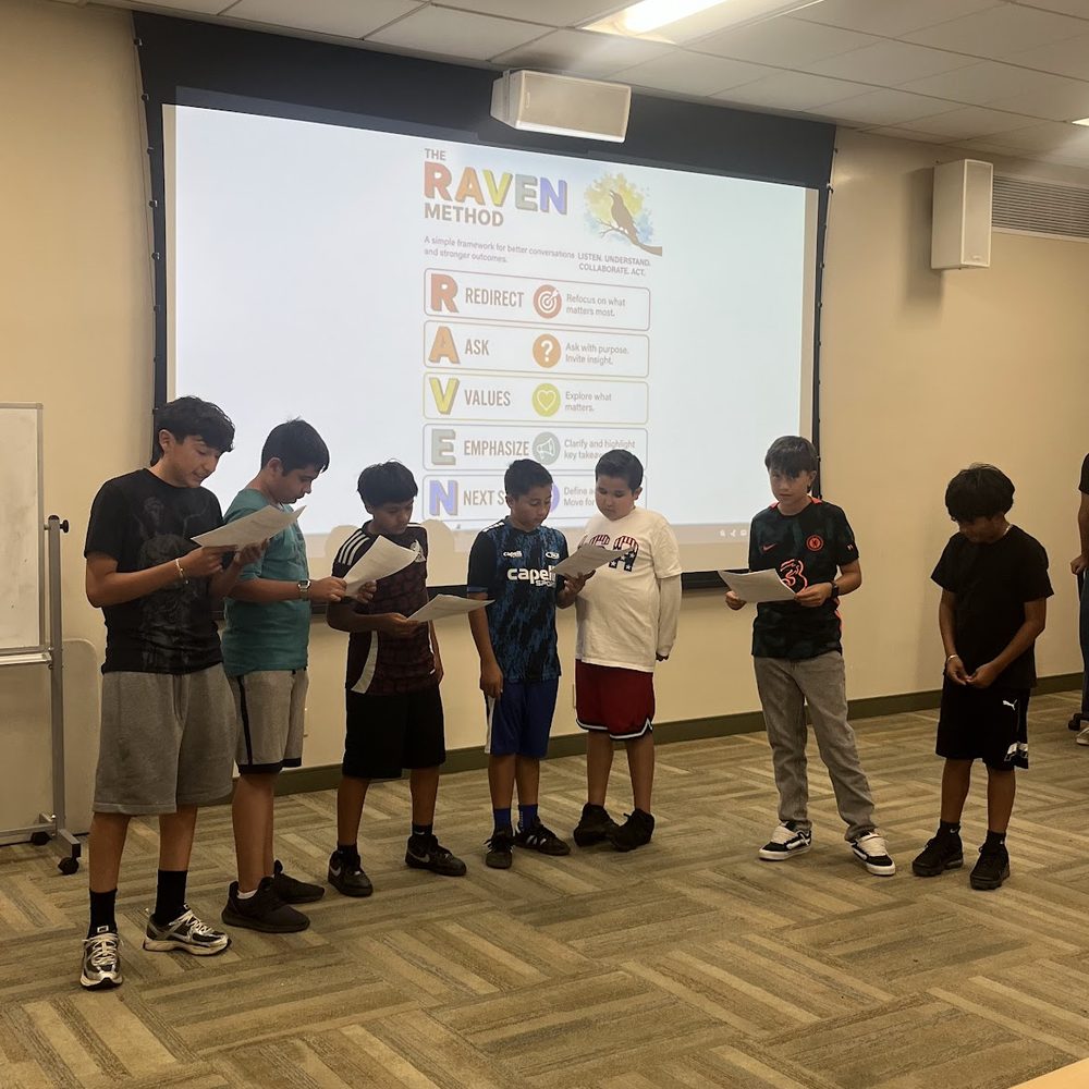 Students standing at the front of a classroom reading from scripts, with the five steps of the RAVEN method projected on the screen behind them.